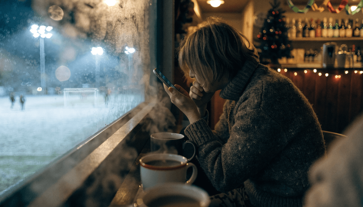 A parent in a woolly jumper sits by a fairy-lit clubhouse window on a December evening holding a smartphone, with a small Christmas tree and a frosty floodlit pitch outside