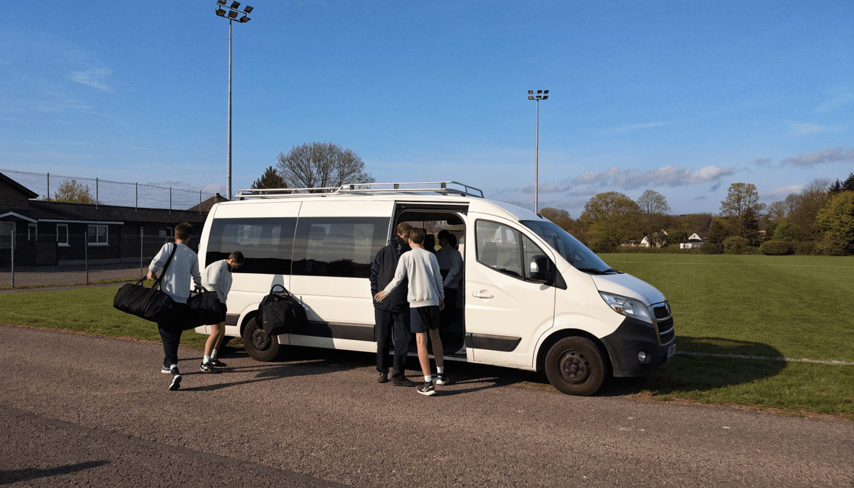 Young players loading kit bags into a white club minibus outside a grassroots sports ground